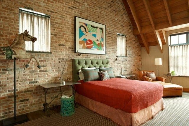 传统床的绿色簇绒床头板靠在有纹理的红砖墙上，墙上装饰着一幅彩色的画，两侧有方形窗帘的窗户。床旁是一张棕色皮革单人沙发椅，与露出木梁的木质天花板相匹配。