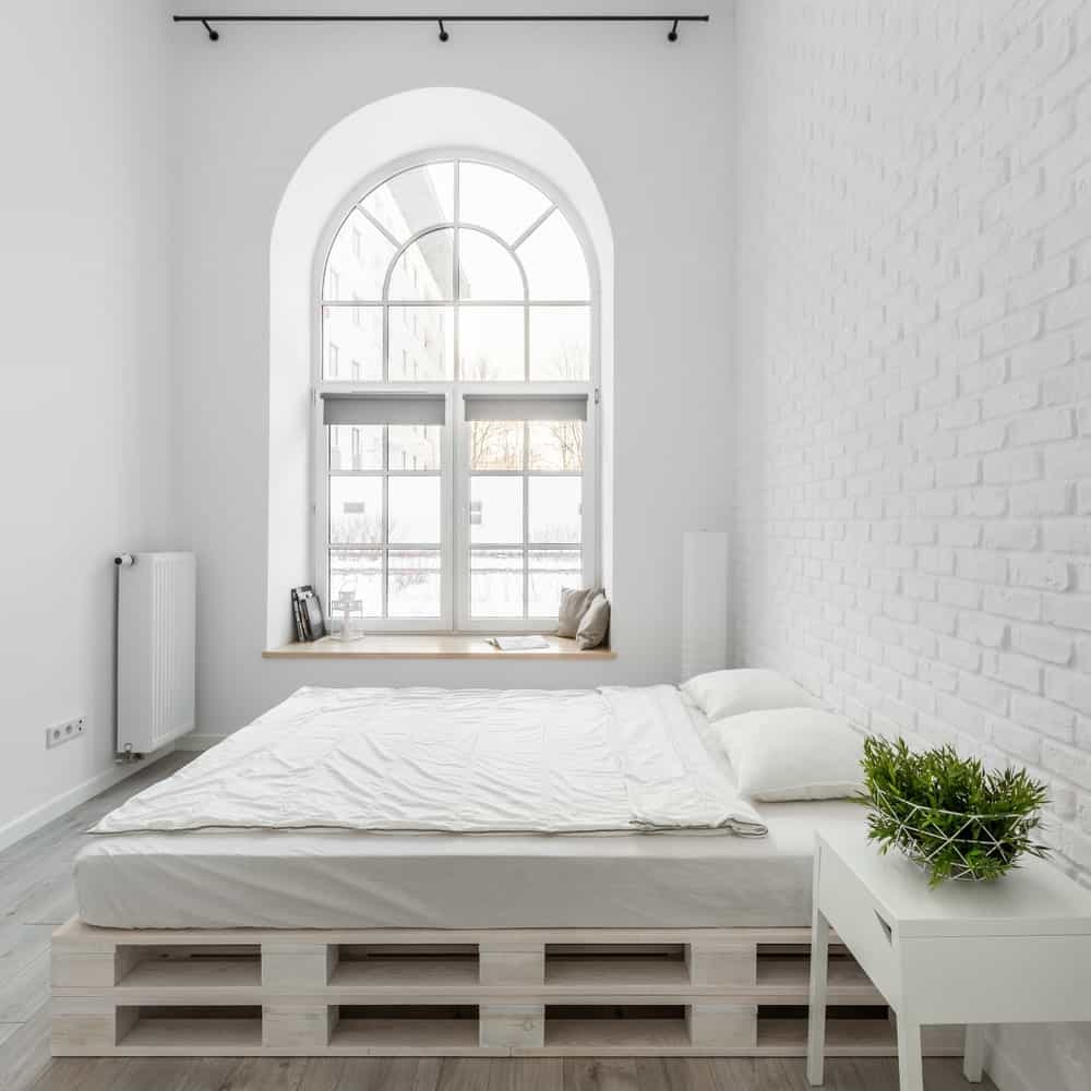 这个工业风格的卧室的亮度来自于高高的拱形窗户，它带来了充足的自然光线，使白色的墙壁和床发光。这张床是在一堆托盘上形成一个平台作为床垫。
