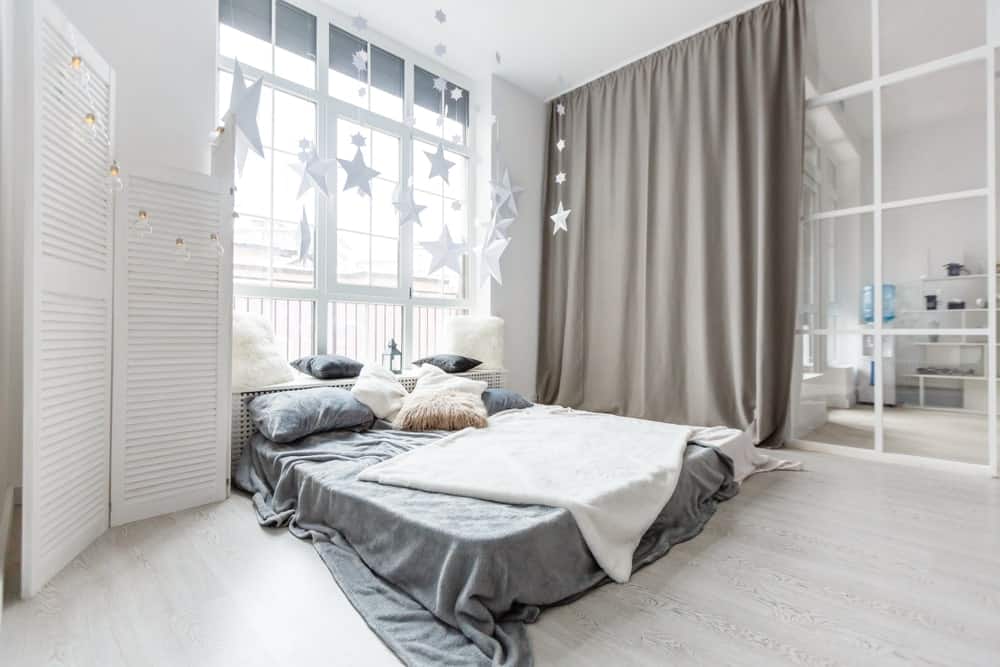 这间工业风格的卧室有高高的白色天花板，浅色硬木地板上放着一张床，看起来更高。床头有一扇高窗，床头有一个木制床头板，也可以作为靠窗的休息区。