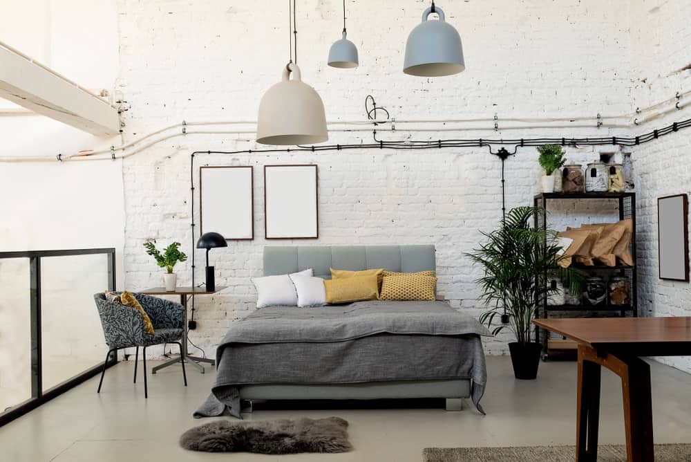 这间工业风格的卧室暴露的电线在白色石材纹理的墙壁上脱颖而出，后面是灰色软垫床头板，与灰色床单相匹配，与浅灰色地板相衬。