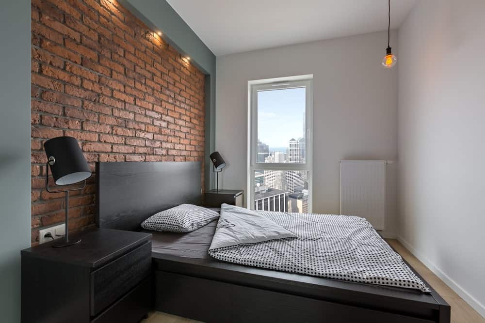 这间工业风格的卧室简洁明了，红色的砖墙与平台式黑色木床的黑色木床头板相得益发。这与床边的黑色木制抽屉和台灯相匹配。
