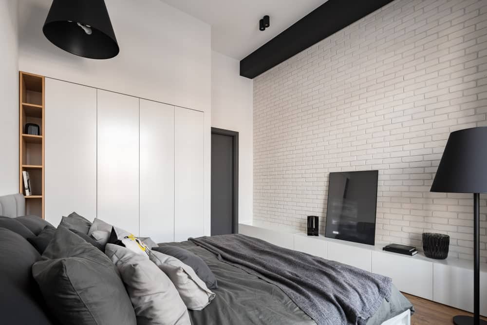 传统床的深灰色床单和枕头与这间以白色为主色调的主卧室相衬，主卧室有白色砖墙，白色橱柜嵌入白墙，白色天花板与黑色吊灯和光束形成对比。