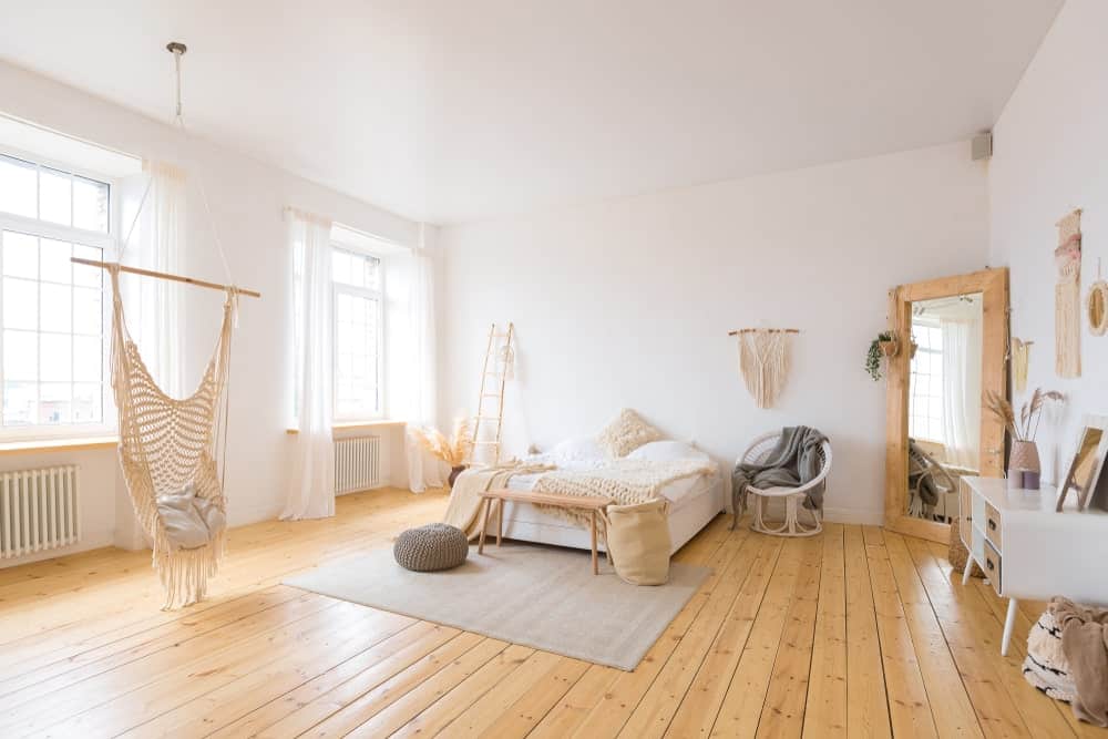 宽敞的破旧别致的主卧室，白色的墙壁和白色的天花板，以及硬木地板。房间提供了一个华丽的床设置和窗户设有可爱的白色窗帘。