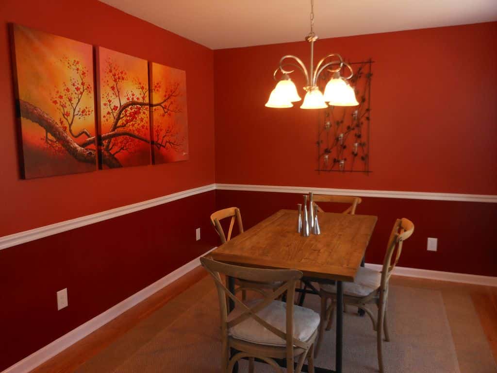 一个不拘一格的红色餐厅，有简单的枝形吊灯，长方形木桌，四把椅子和苦恼的靠垫，地毯，绘画，锻铁展示板，和金属中心装饰品。