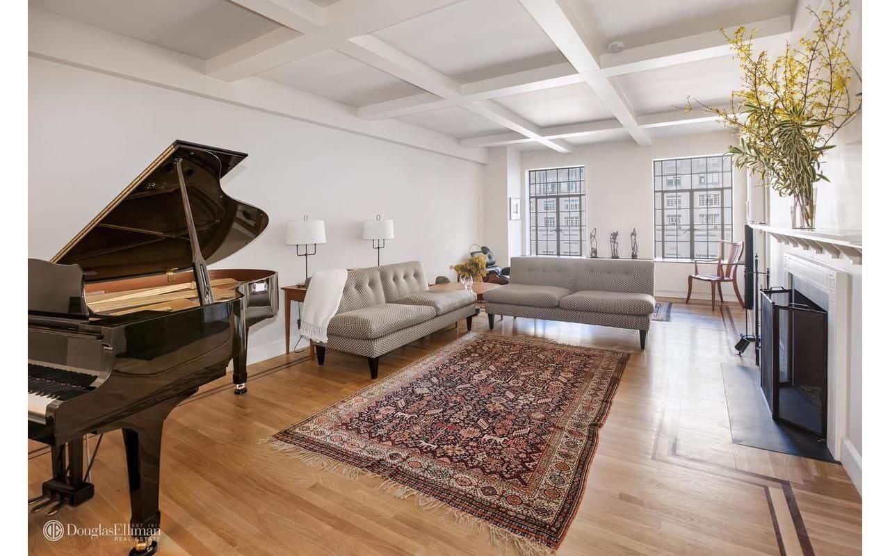 白色的客厅里有灰色的簇绒沙发、一架小型三角钢琴和一座覆盖着金属栅栏的白色壁炉。它有格子天花板和硬木地板，上面铺着复古地毯。
