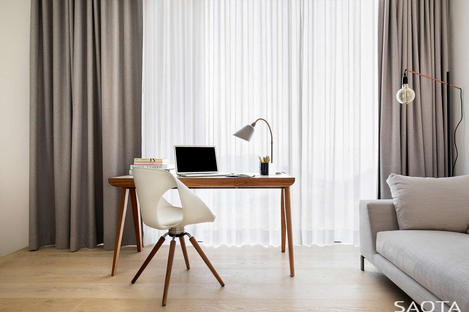 家庭办公室的通高玻璃窗上覆盖着灰褐色的窗帘和白色的透明窗帘。它有一张木制办公桌，还有一把时髦的白色椅子，底座像木制的爪。