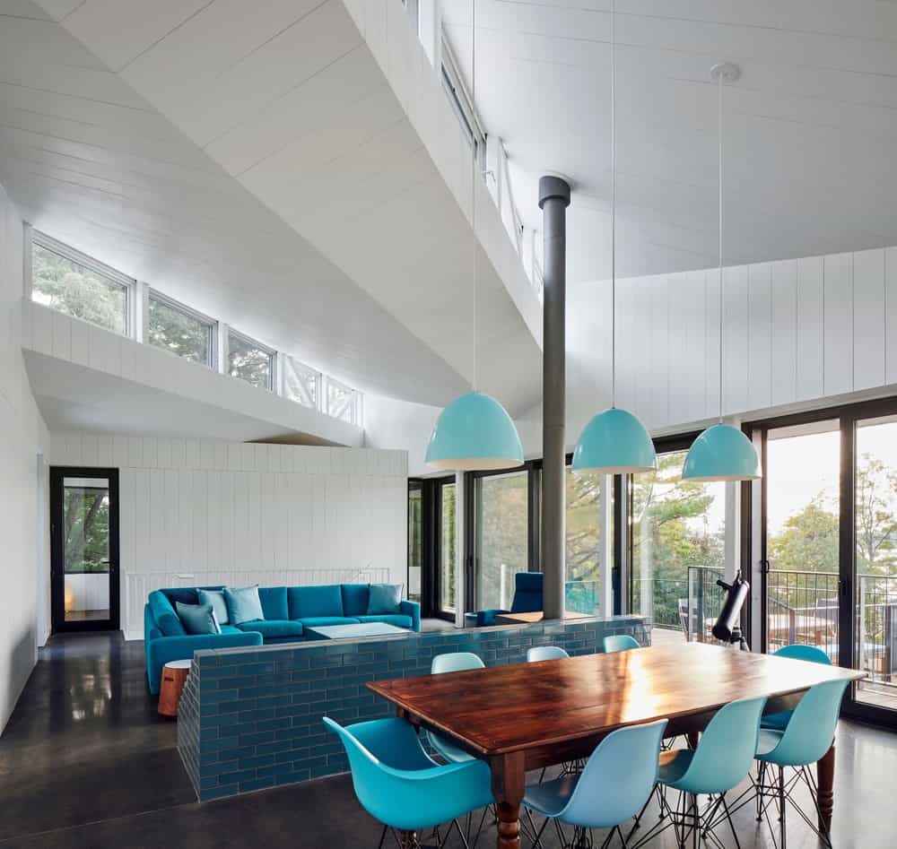 白色的墙和天花板用蓝色的组合板、砖分隔板、天蓝色的吊灯和围绕着长方形木桌的餐椅进行装饰。