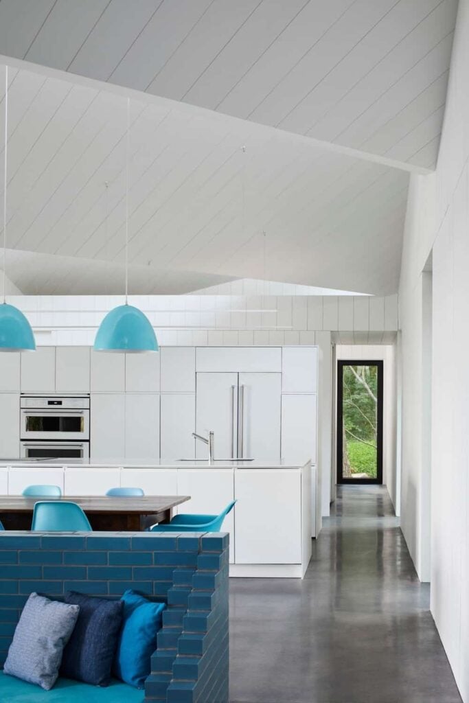 现代风格的白色内餐厨房，蓝色圆顶吊坠悬挂在船板天花板上，木制餐桌周围环绕着现代风格的椅子。