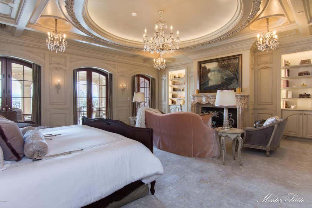 豪华的主卧室，有一张木床和壁炉旁的座位区，由华丽的圆形托盘天花板上悬挂的花哨水晶吊灯照明。