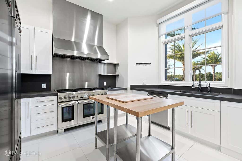 白色的现代厨房，配有不锈钢电器和后挡板，两侧是黑色的后挡板和台面，窗户，以及一个小的不锈钢餐桌厨房岛，带轮子和木质表面，位于白色瓷砖地板上。