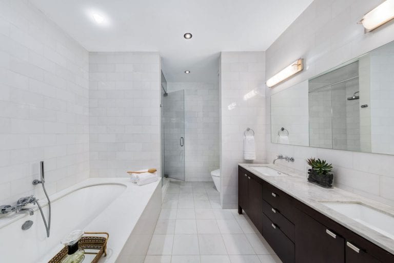 白色浴室拥有一个长浴缸和一个带双水槽的步入式淋浴，配有大理石台面，由壁灯照明。