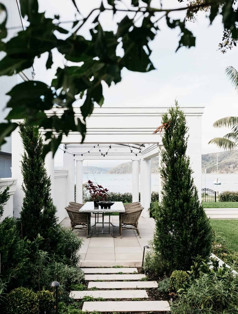 与树和绿色植物围拢的美丽的通路的白色露台导致室外餐桌集合的。