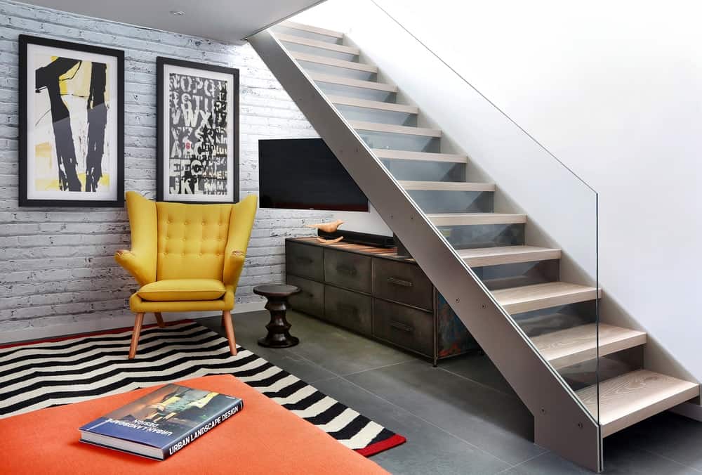 配有无框玻璃栏杆和轻质木踏板的现代楼梯。它下面有一个乡村风格的抽屉，一张黄色的簇绒椅子和圆形的边桌，上面铺着黑色条纹和橙色分层的地毯。