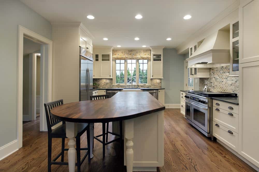 厨房有浅蓝色的墙壁，白色的橱柜和装饰物，木地板，以及一个l形的厨房岛，有木质表面和座椅。