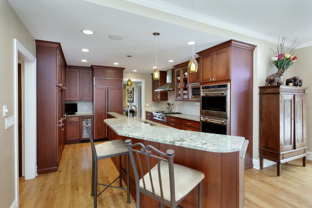 这个厨房有一个棕色的橱柜和一个狭窄的厨房岛，带一个大理石台面，由吊灯和嵌入式灯照亮。