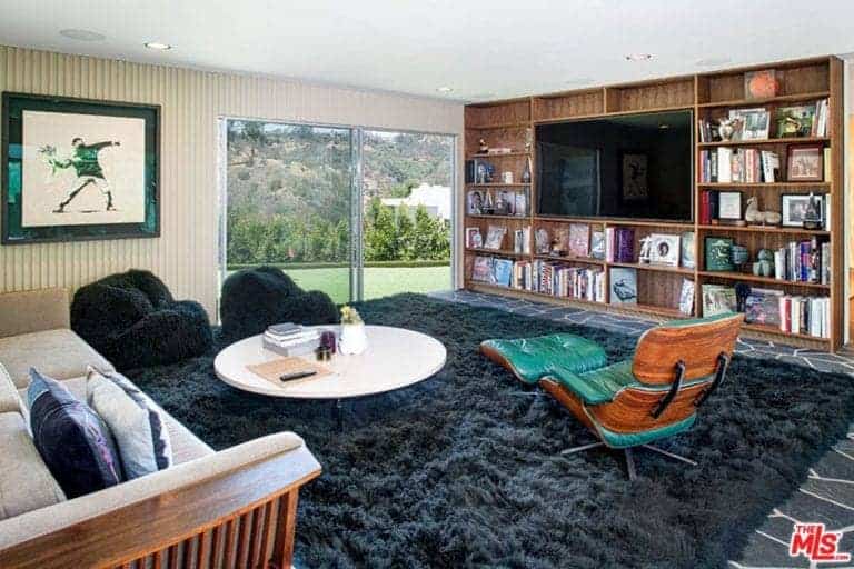 一个平板电视安装在客厅的内置架子上，装饰有墙壁艺术，安装在床头板墙上。它包括一个米色沙发和圆形咖啡桌，还有黑色舒适的椅子和一张绿色的躺椅，上面是蓬松的地毯。
