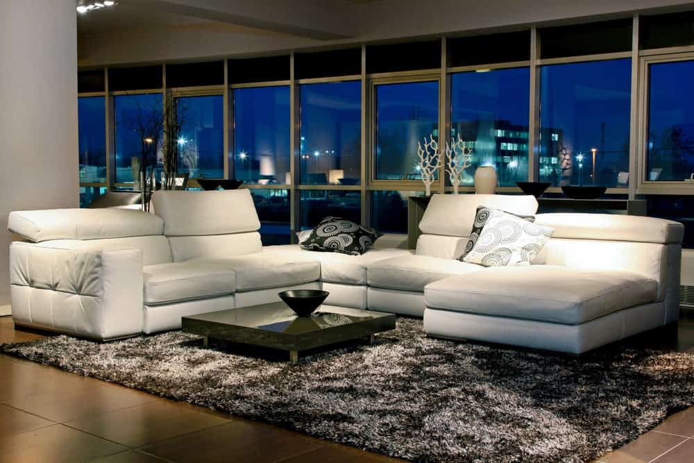 宽敞的现代客厅，瓷砖地板，上面有时尚的地毯，白色沙发和小中心桌。该区域被玻璃墙和窗户包围。