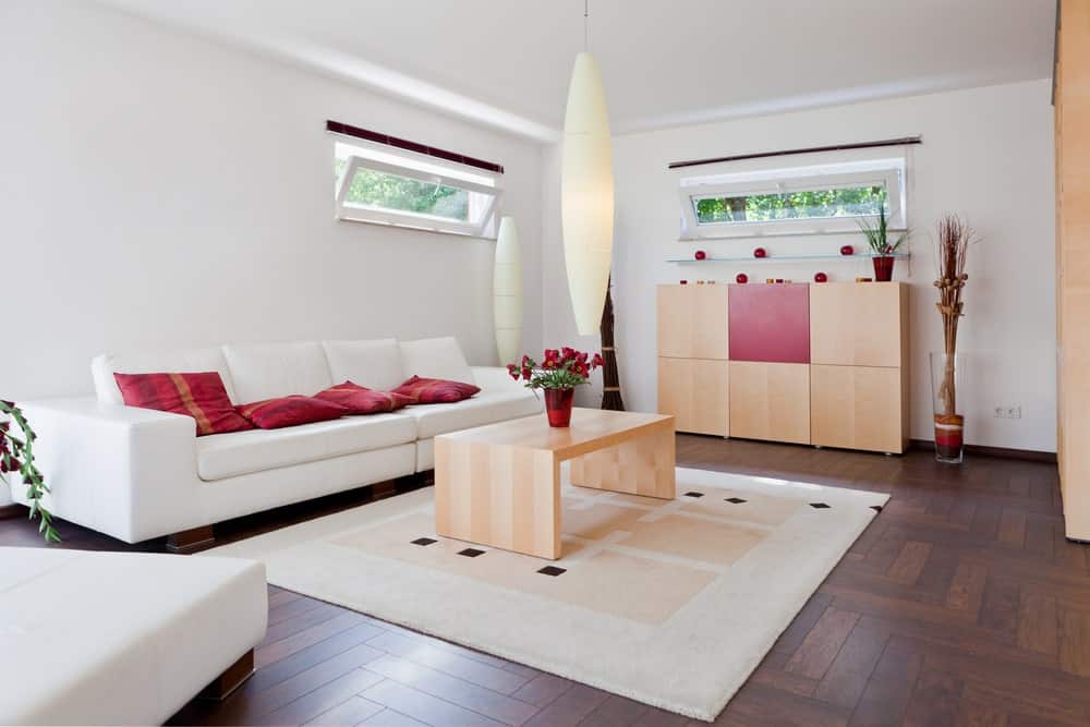 华丽的客厅，白墙，硬木地板，上面铺着小地毯。房间里有一张木制的中央餐桌和橱柜，还有一张白色沙发和红色抱枕。
