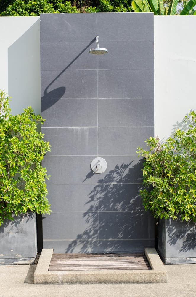 现代户外淋浴，灰色墙壁和高度设置的淋浴头被绿色植物包围。