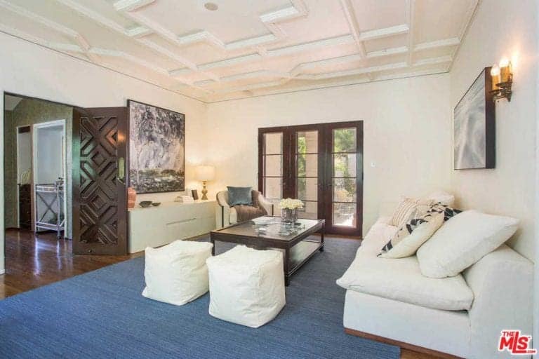 一扇雕花木门通往这间客厅，客厅里有白色沙发和凳子，它们挨着深色木咖啡桌，上面铺着蓝色地毯。