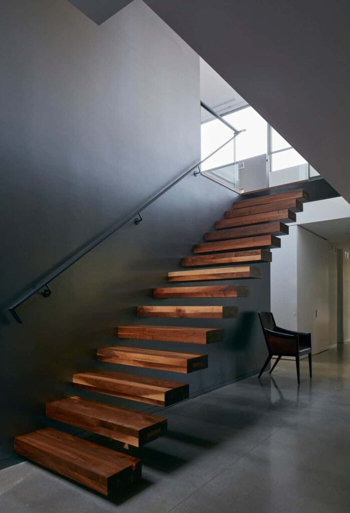 直的浮动楼梯，木制踏板和黑色钢扶手固定在灰色的墙壁上。铺着瓷砖的地板下面有把椅子。