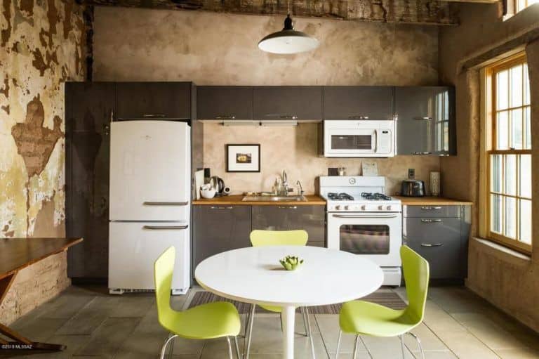质朴的餐厅厨房展示了灰色光滑的橱柜和白色电器，以及圆形餐桌和由圆顶吊坠照明的绿色现代椅子。