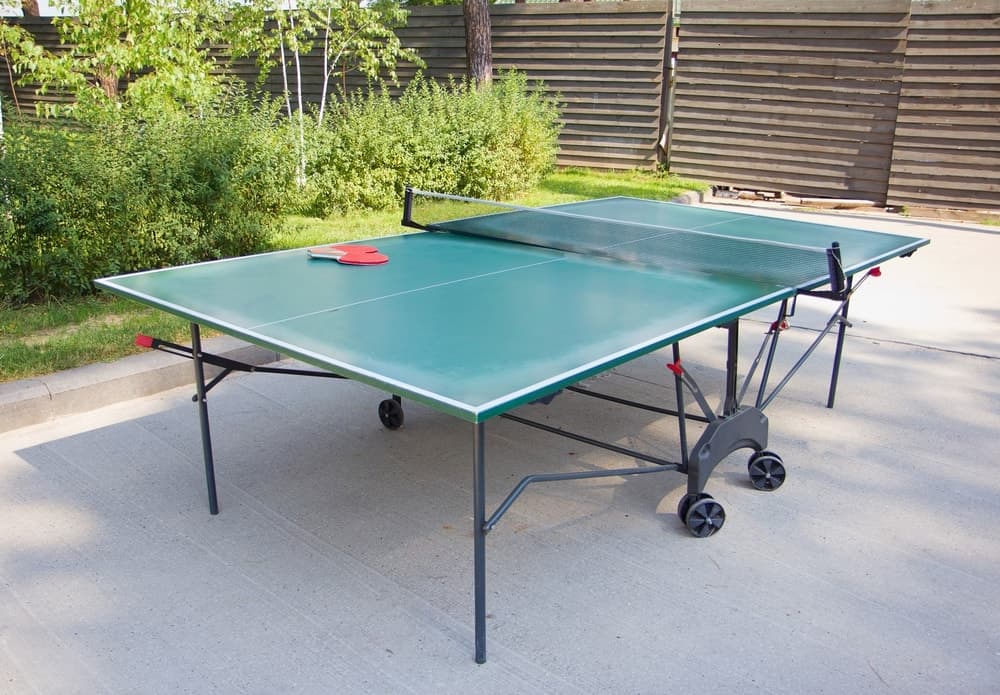 绿色室外乒乓球桌，轮子放在室外的混凝土地板上，附近有木制私人栅栏和绿色植物。