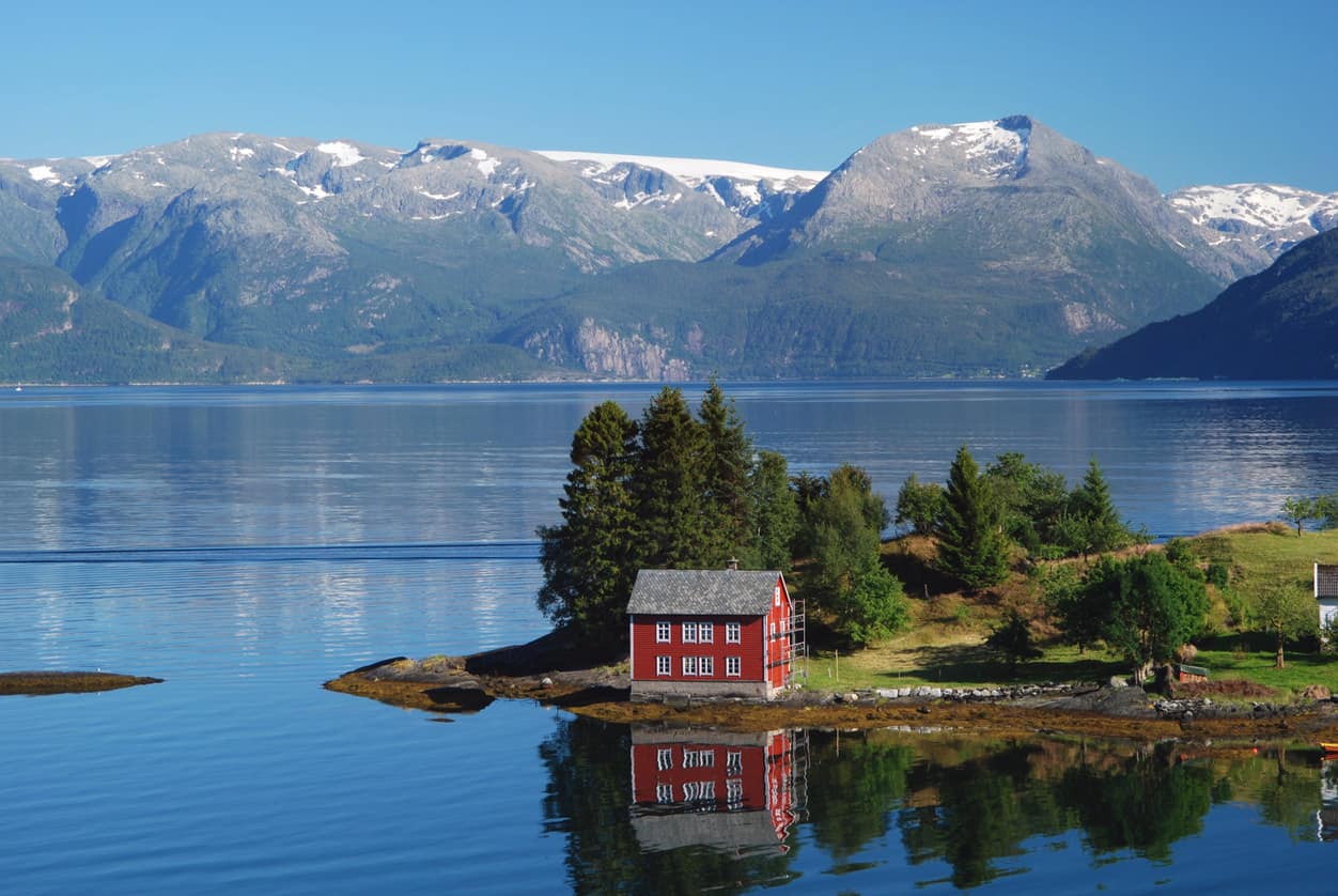 夏天在挪威西南部的哈当厄峡湾。一座红色的挪威房屋坐落在峡湾的一个小岛上。远处是Folgefonna冰川。