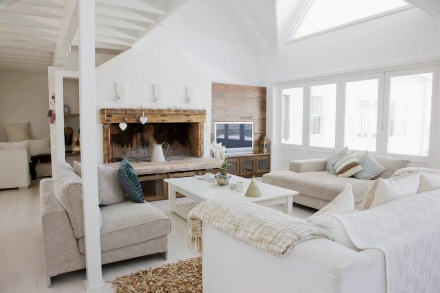 这是一间淡雅质朴的客厅，有高耸的白色天花板和破旧的横梁壁炉架。浅米色的沙发与原本全白的空间搭配在一起。
