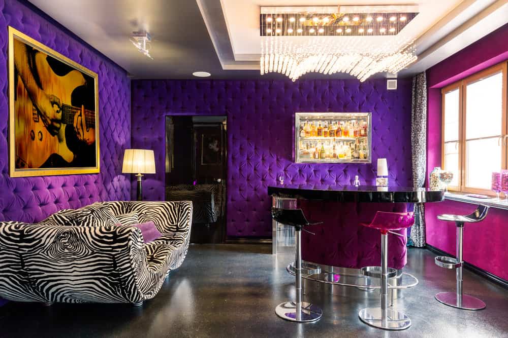 这是一个完全迷人的家庭酒吧和休息室，有紫色的簇绒墙，紫红色和铬色的凳子，粉红色的窗户墙，黑色的地板和一个超级野生斑马沙发。还有，看看那盏不可思议的吊灯。
