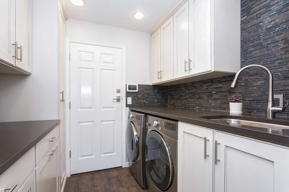 现代洗衣房设有白色橱柜，硬木地板，实用水槽，黑色台面，和配套的黑色，前置洗衣机和烘干机。
