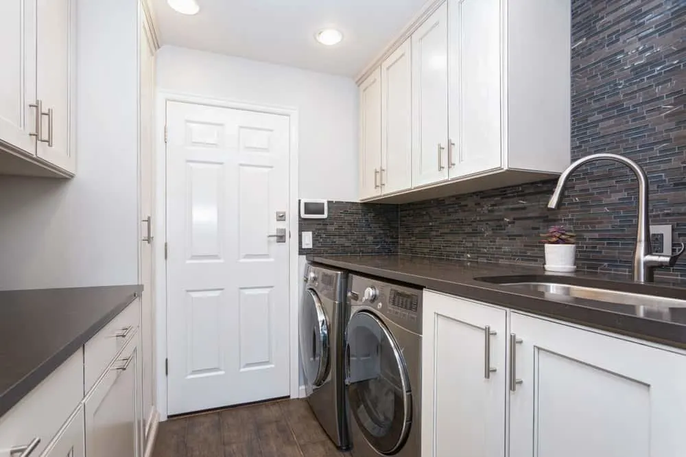 当代洗衣房特性白色橱柜、实木地板、效用水槽,黑色的台面,搭配黑色,前置式洗衣机和干衣机。