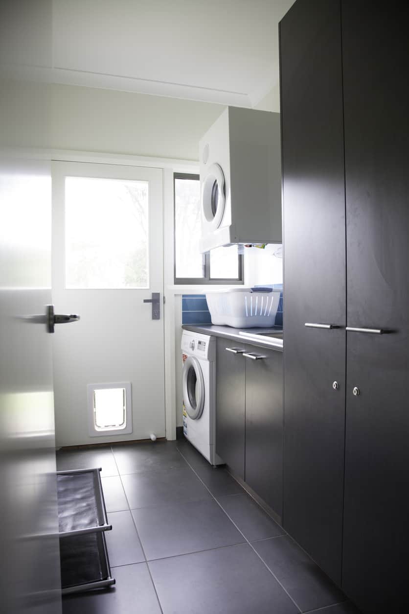 小型洗衣房设有洗衣机上方的壁挂式烘干机。这个房间还包括深灰色的储物柜、瓷砖地板和一扇宠物门，作为通往室外的外门的一部分。