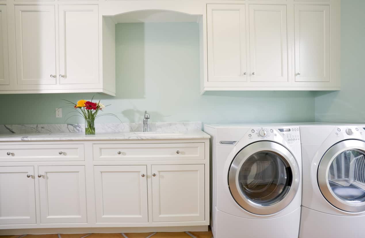 洗衣房设有定制白色橱柜，背靠浅绿色墙壁，前置洗衣机和烘干机组和瓷砖地板。
