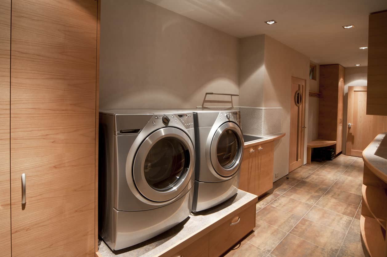 白色和深灰色的现代洗衣房设有宽敞的存储空间，轻质木地板，前置洗衣机和烘干机，以及从侧窗和后门射入的大量自然光。