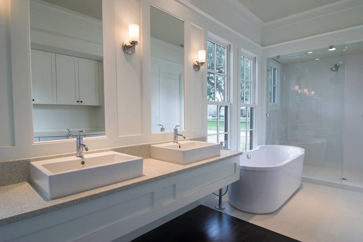 长长的白色主浴室，有独立的白色浴缸和大型步入式淋浴。包括长梳妆台和两个现代风格的水槽。浴缸旁边的高窗。还有，看看地板，一部分是瓷砖，另一部分是深色木材。