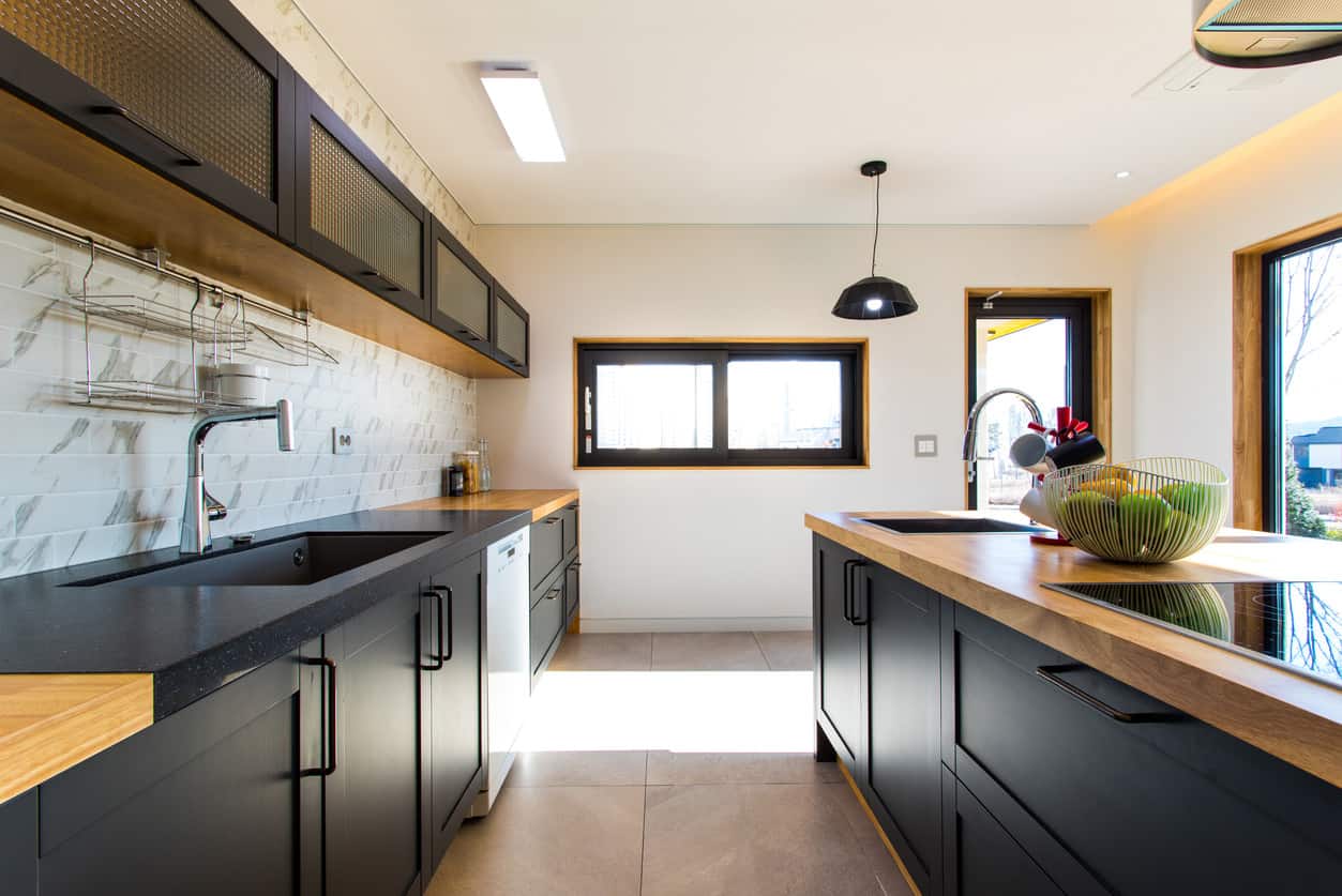 浅木和黑色构成了这个现代厨房的配色方案。这是一面墙，有着长岛的布局。这是一个非常简单的美学和相当明亮的结果，这是由于窗户，白色的后挡板和白色的天花板。
