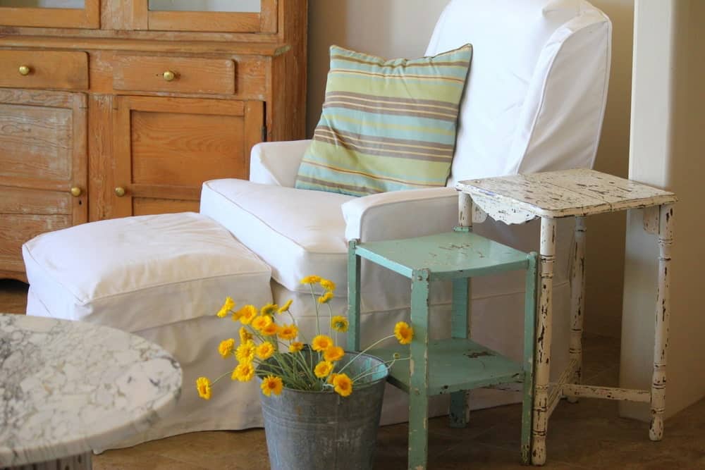一张躺椅和一张铺着白色沙发套、带抱枕的软凳，旁边是一对涂过漆的边桌和一桶花。