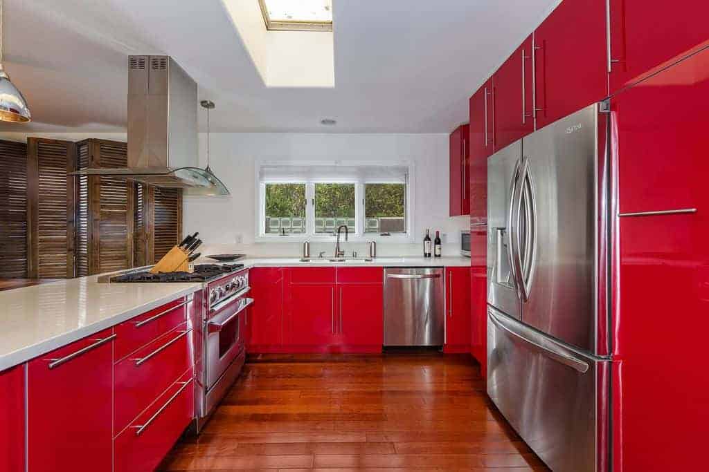 大u型厨房，明亮的红色橱柜和不锈钢器具，白色台面和白色天花板。我认为红色的木地板没有提供足够的对比。我想浅色的木地板会更好看。