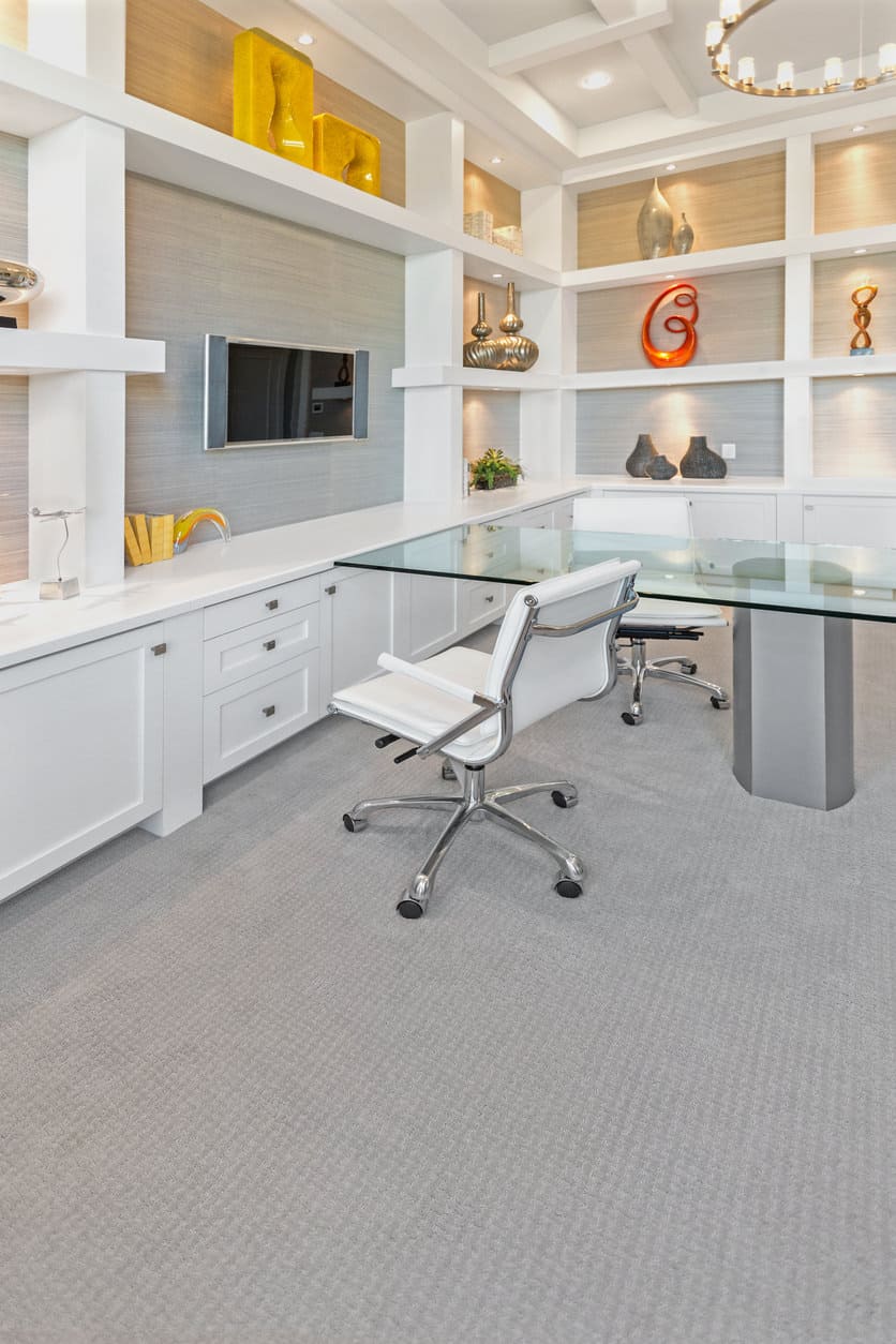 这是一间很大的家庭办公室，铺着灰色的地毯地板，有白色的橱柜、架子和柜台，还有一张时髦的玻璃桌面、格子天花板和一盏令人惊叹的枝形吊灯。