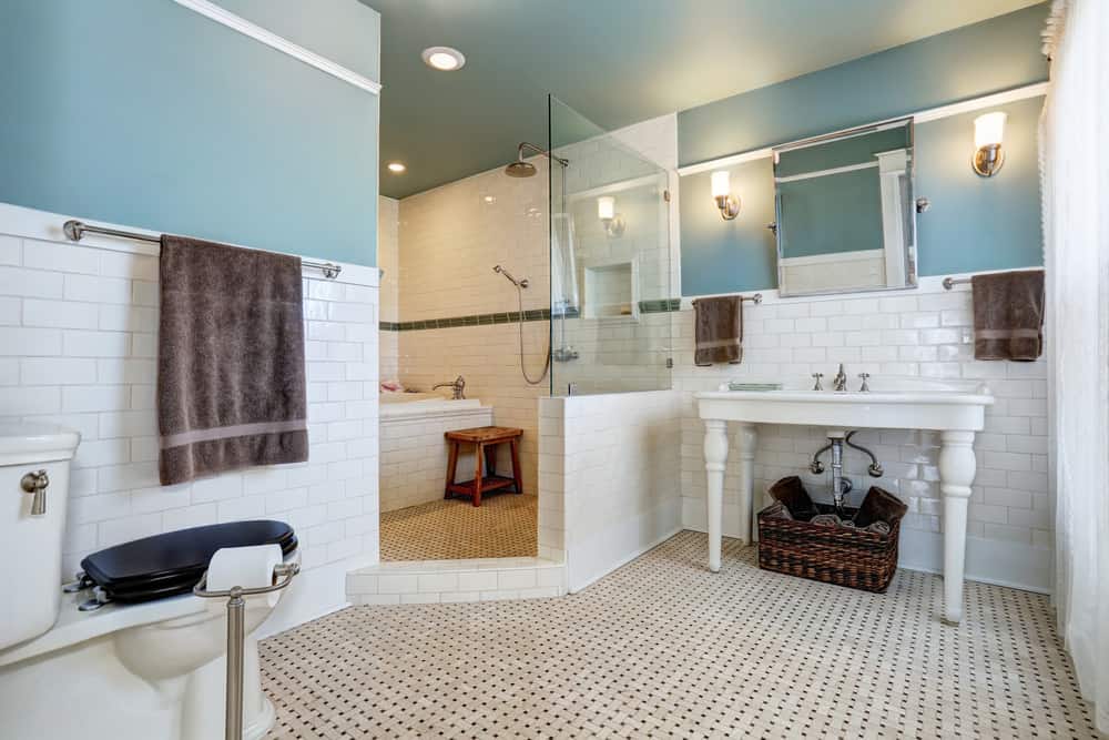 这个浴室融合了古老与现代，城市与乡村。房间里有漂亮的淡蓝色墙壁，白色瓷砖和深蓝色的马桶盖，与浴室旁边的木制踏步凳和水槽下面的柳条篮子形成了鲜明的对比。