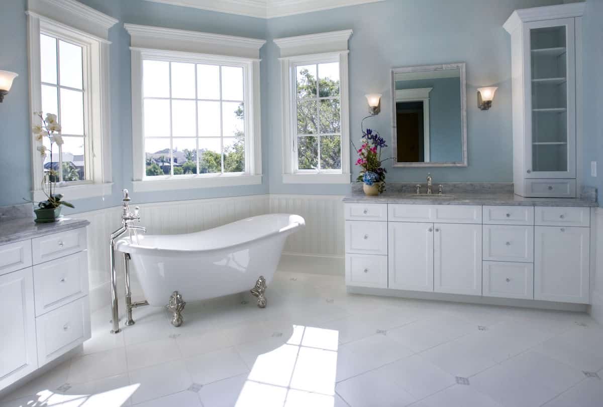 这个浴室非常女性化，它的淡蓝色墙壁和毛巾柜顶部的许多浮华。从浴缸上方打开的窗户射进来的自然光加强了柔和的阴影。