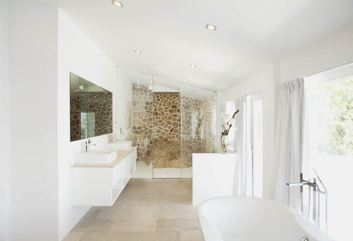 这个主浴室比大多数典型的住宅厨房更宽敞，当它涉及到一个真正放松的淋浴和沐浴体验时，是快乐的缩影。墙壁上安装的白色梳妆台和棕褐色的台面与质朴的石头图案淋浴墙和岩石形状的淋浴座相呼应。透明的玻璃镶板和独立的现代风格花园浴缸在阳光下闪闪发光，穿过三扇大窗户。