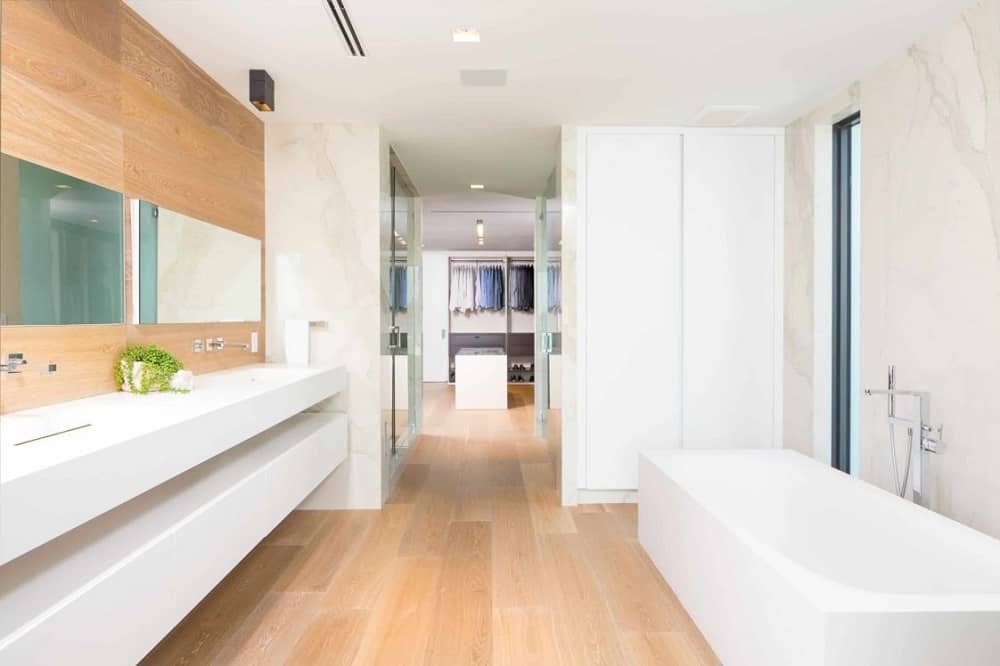 这是一间浴室，在白色浴缸的对面，有一个现代的白色漂浮梳妆台，在硬木地板的映衬下，与梳妆台镜子后面的墙壁相匹配。