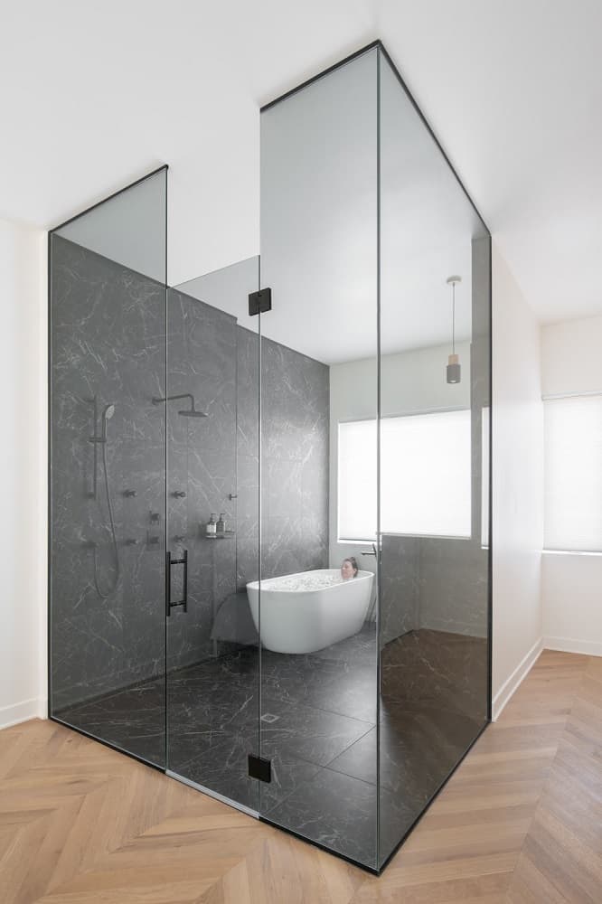 这是一个近距离观察的浴室，它的两面都有玻璃墙，一面是黑色大理石，它的地板使远处角落的白色独立浴缸脱颖而出。