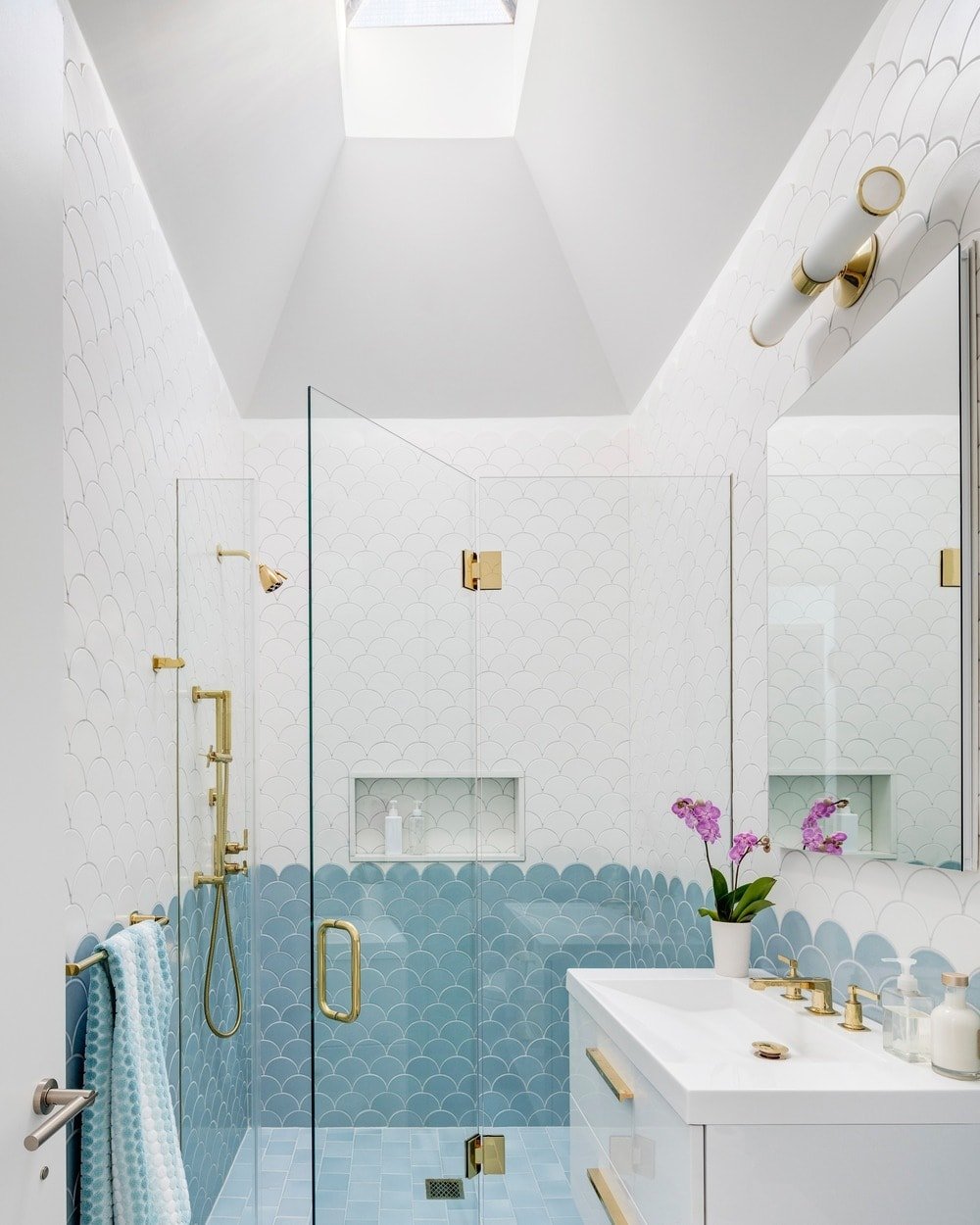 这个简单的浴室的墙上贴着瓷砖，从上部墙壁的白色瓷砖到齐腰高的浅蓝色瓷砖，看起来像鱼鳞。这给人一种美丽的锦鲤鱼的印象，浴室装置上的少数金色口音在玻璃和白色元素的映衬下更加突出。