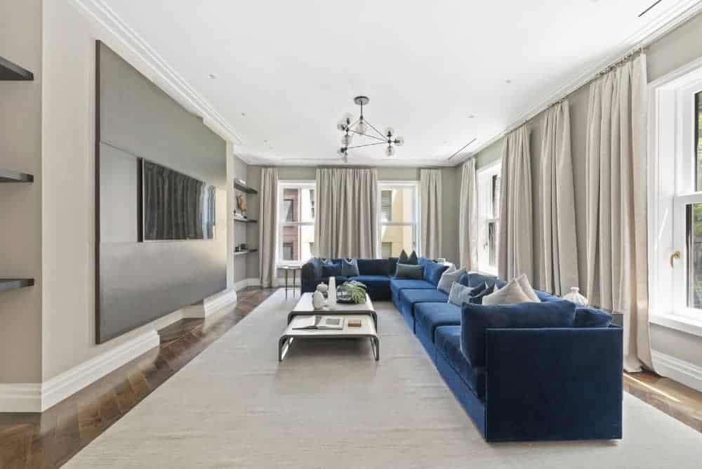 蓝色天鹅绒截面脱颖而出在这个现代家庭房间,灰色的墙壁,平板电视,拼花地板和模块化的咖啡桌米色地毯。现代全球玻璃吊灯完成了。
