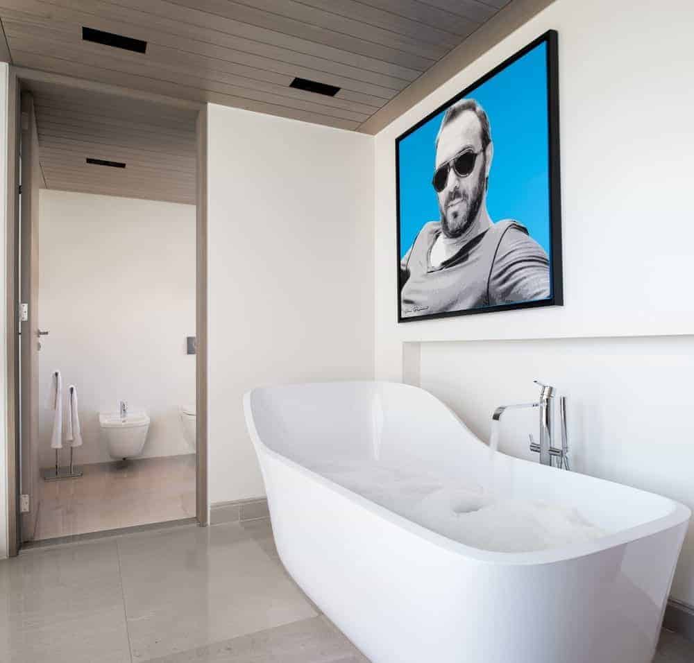 一幅巨大的肖像为这个现代的主浴室增添了一个很好的口音，在瓷砖地板上有一个独立的浴缸，配有铬固定装置。房间一侧的木门通向厕所区域。