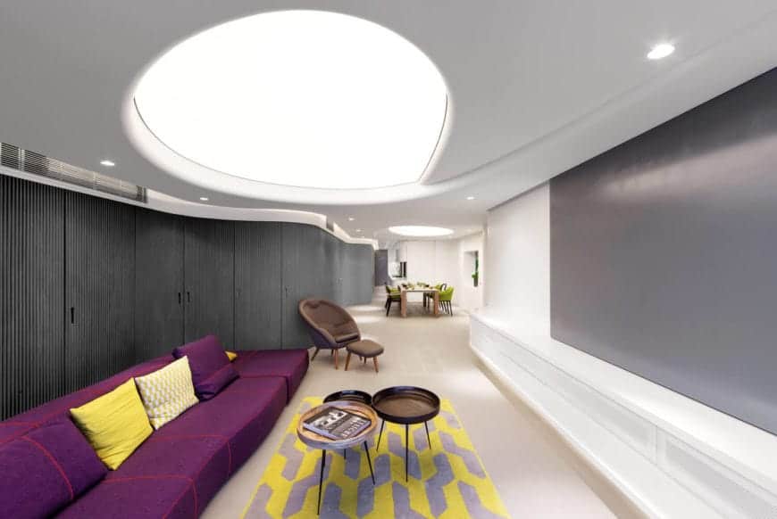 圆形凹形天花板，加上嵌入式照明，在这个时尚的现代客厅中做出了大胆的陈述。紫色的长沙发，配上黄色的靠枕，与房间其他地方的中性色形成了鲜明的对比。