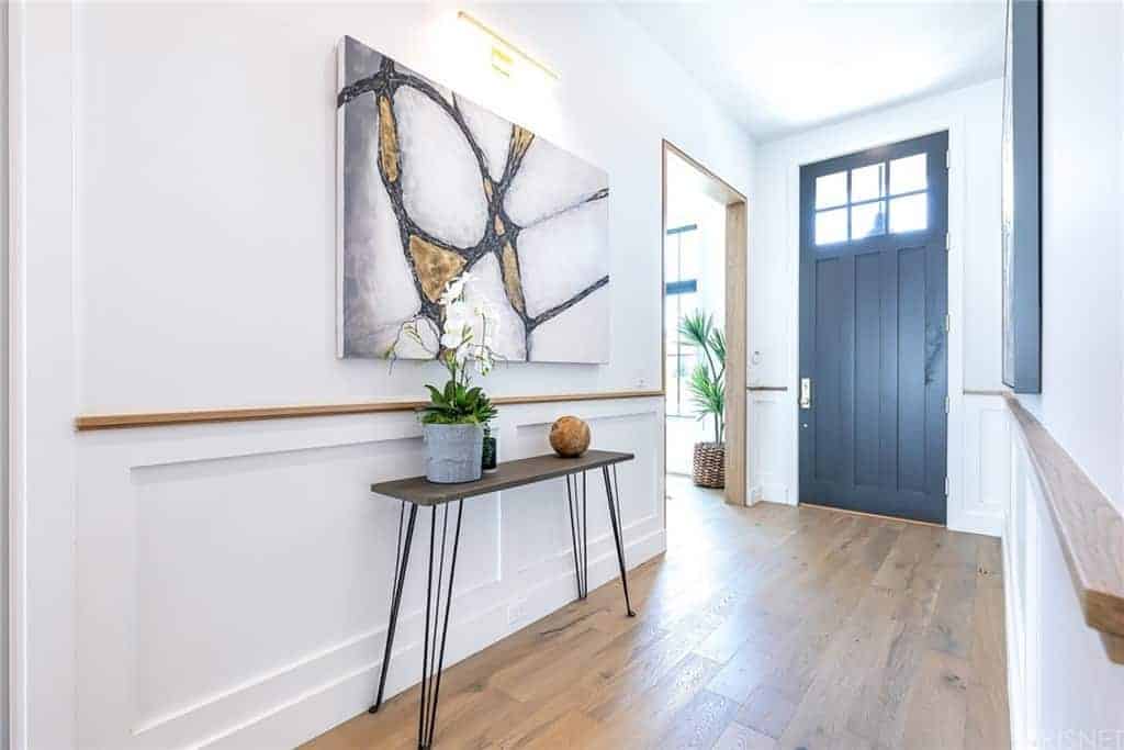 蓝灰色工匠风格的前门通向明亮的白色门厅，门厅设有护墙板、硬木地板，以及一幅当代抽象画下面的木顶控制台。